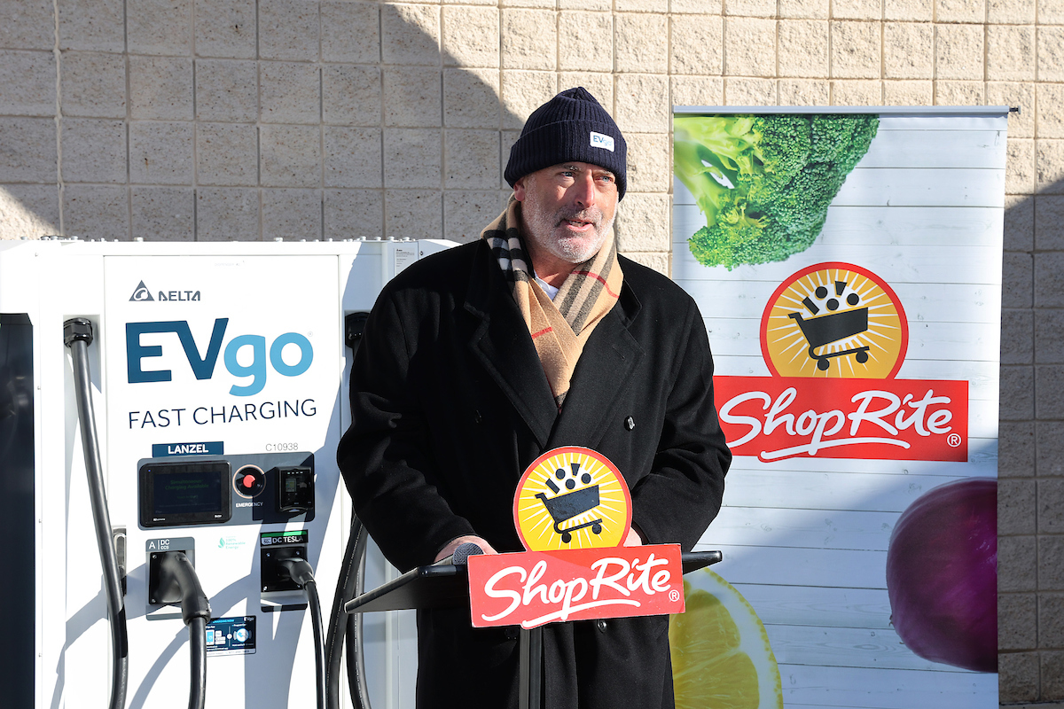 A man stands near an EVGO fastcharging station holding a ShopRite sign. He is sporting a beanie and a scarf, and has a trimmed white beard. To his right, is a ShopRite sign with a shopping cart and rays of sunshine showing behind it.
